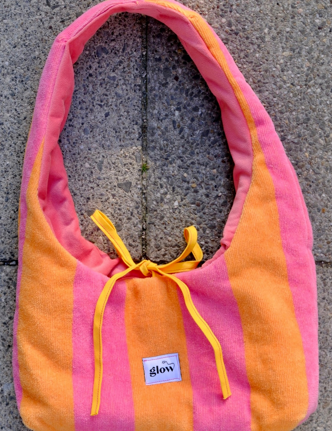 A pink and orange striped bath towel shoulder bag with yellow drawstrings and a small white 'glow' brand label, laid flat on a gray stone surface.