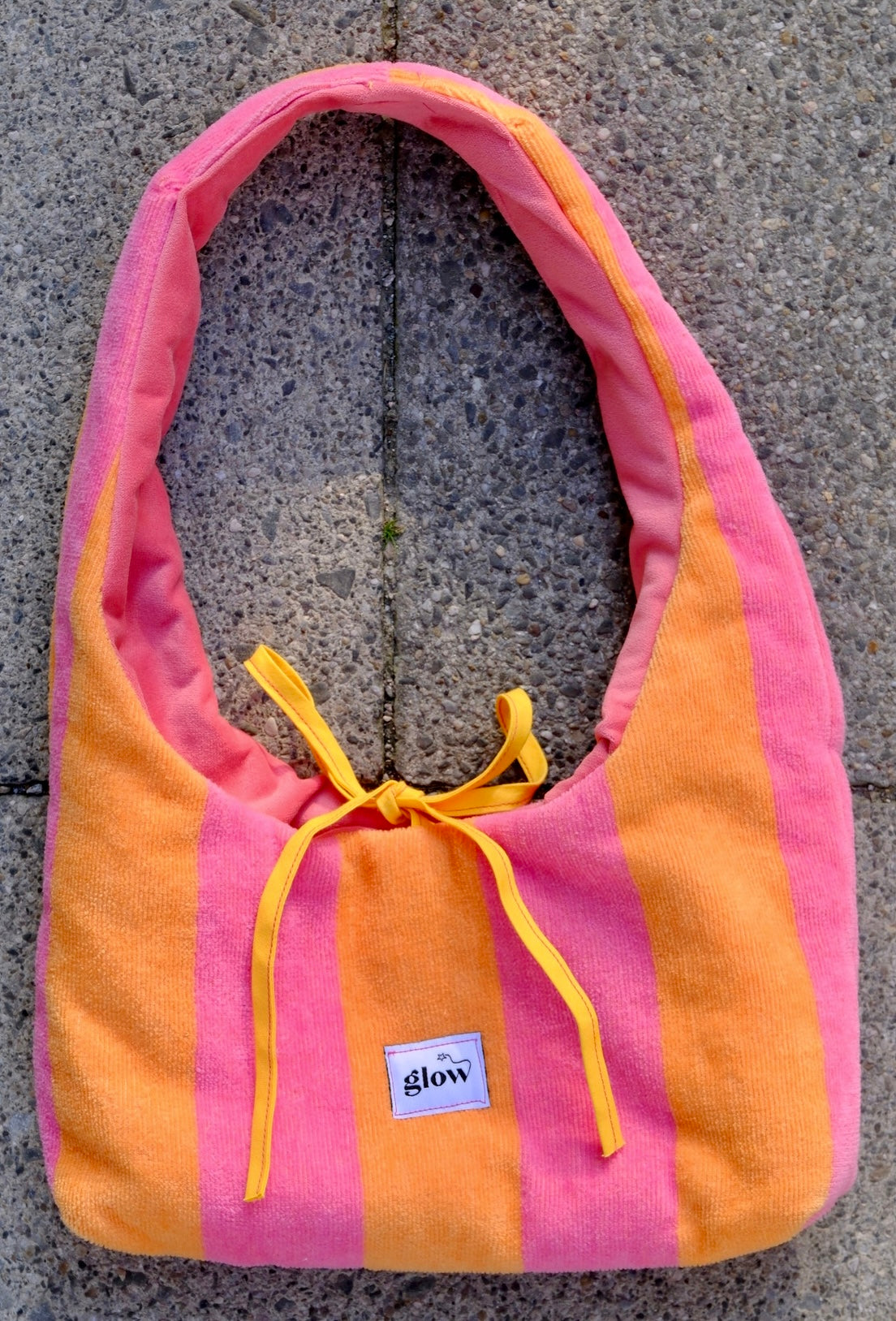A pink and orange striped bath towel shoulder bag with yellow drawstrings and a small white 'glow' brand label, laid flat on a gray stone surface.
