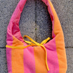 A soft fabric shoulder bag with vertical pink and orange stripes and a yellow drawstring tie, laid flat on a gray concrete surface.