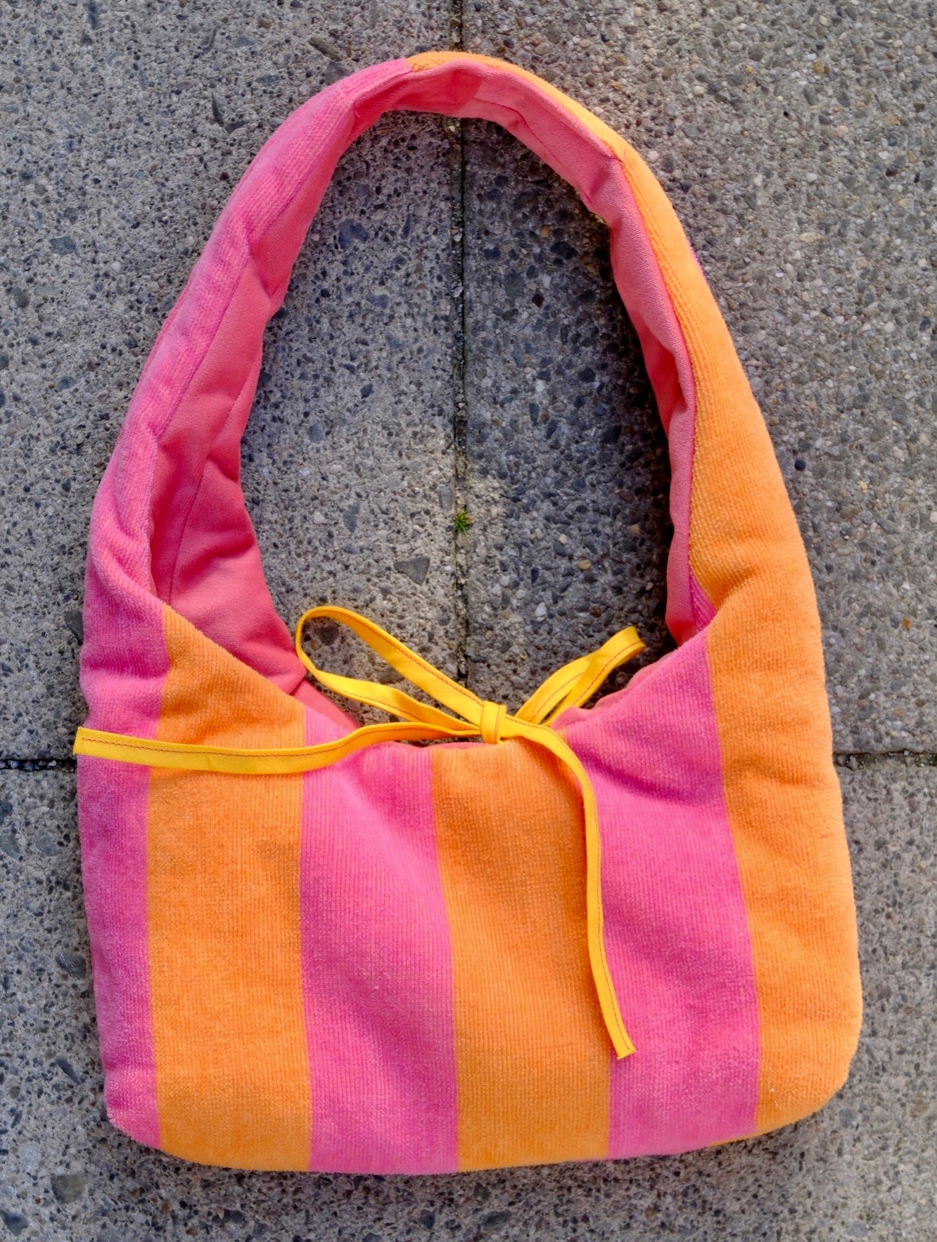 A soft fabric shoulder bag with vertical pink and orange stripes and a yellow drawstring tie, laid flat on a gray concrete surface.