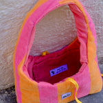 A pink and orange striped plush dog bed with a yellow drawstring, photographed outdoors against a textured beige wall and gray pavement. The brand name 'stixx' is visible on a small white label.