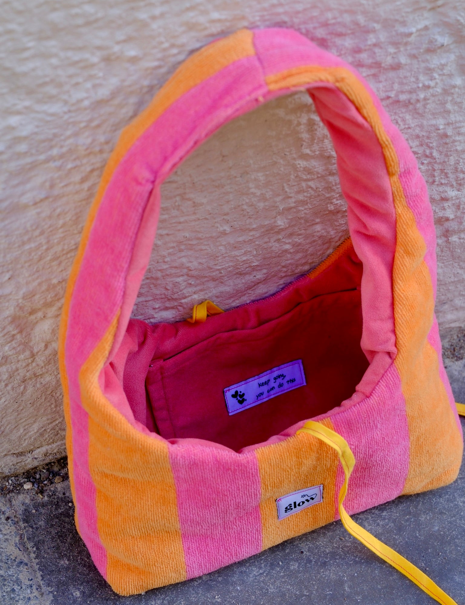 A pink and orange striped plush dog bed with a yellow drawstring, photographed outdoors against a textured beige wall and gray pavement. The brand name 'stixx' is visible on a small white label.