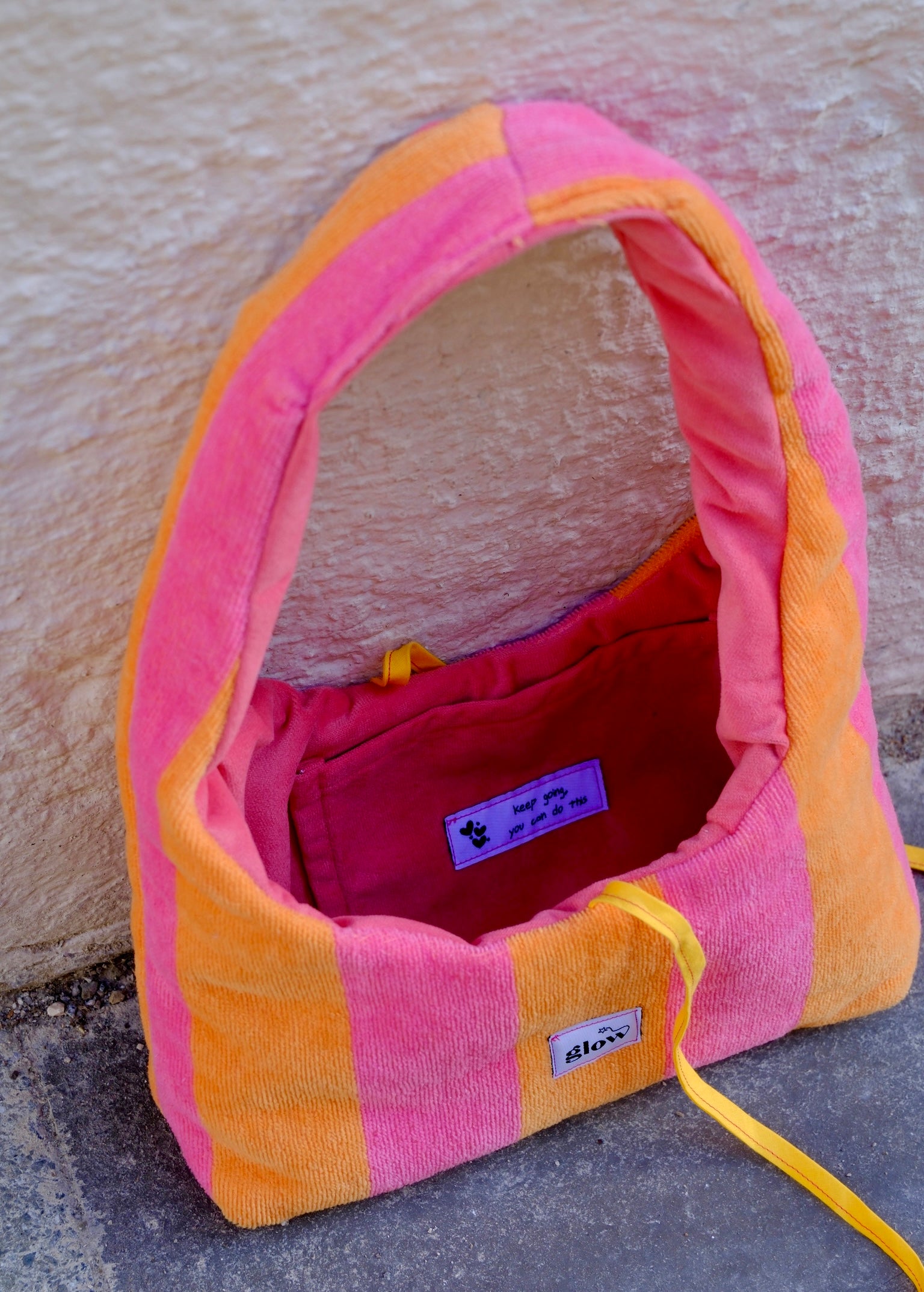 A pink and orange striped plush dog bed with a yellow drawstring, photographed outdoors against a textured beige wall and gray pavement. The brand name 'stixx' is visible on a small white label.