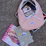 A light pink corduroy shoulder bag with a white drawstring and a visible 'glow' brand patch, shown open with various items inside on a gray asphalt surface next to a magazine and a decorated notebook.