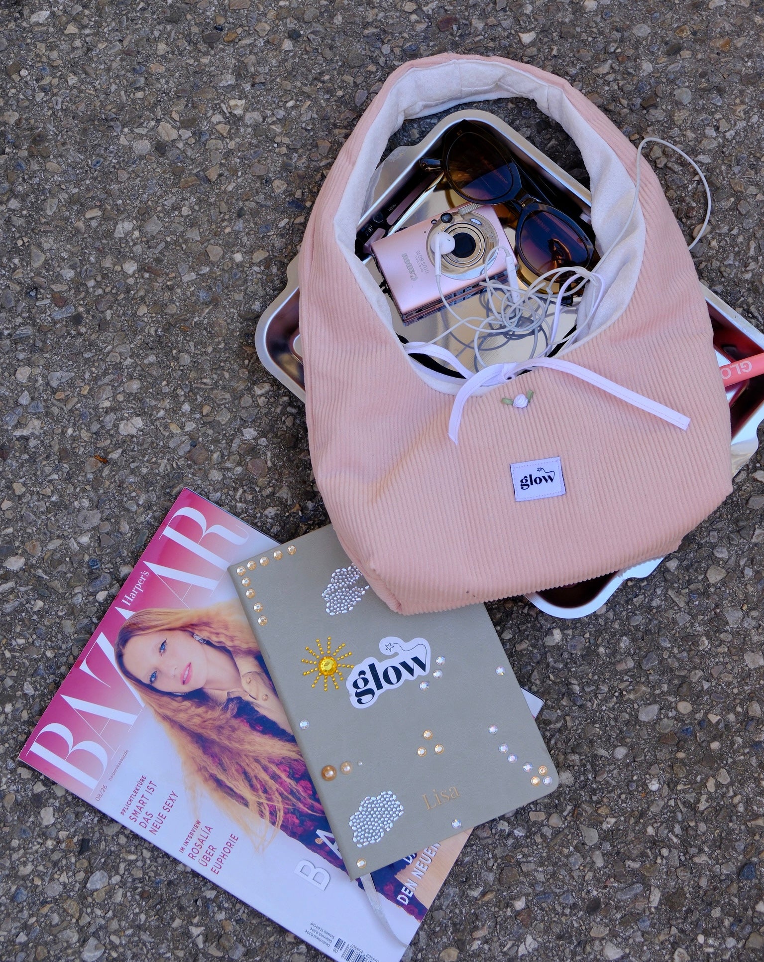 A light pink corduroy shoulder bag with a white drawstring and a visible 'glow' brand patch, shown open with various items inside on a gray asphalt surface next to a magazine and a decorated notebook.