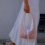 A person wearing a white tiered maxi skirt and a light blue striped top holds a dusty pink slouchy shoulder bag with a white bow detail. The person is wearing brown flip-flop sandals and standing outdoors on a stone floor.
