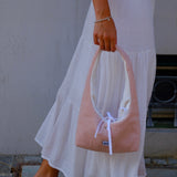 A person wearing a white tiered maxi skirt and a light blue striped top holds a dusty pink slouchy shoulder bag with a white bow detail. The person is wearing brown flip-flop sandals and standing outdoors on a stone floor.