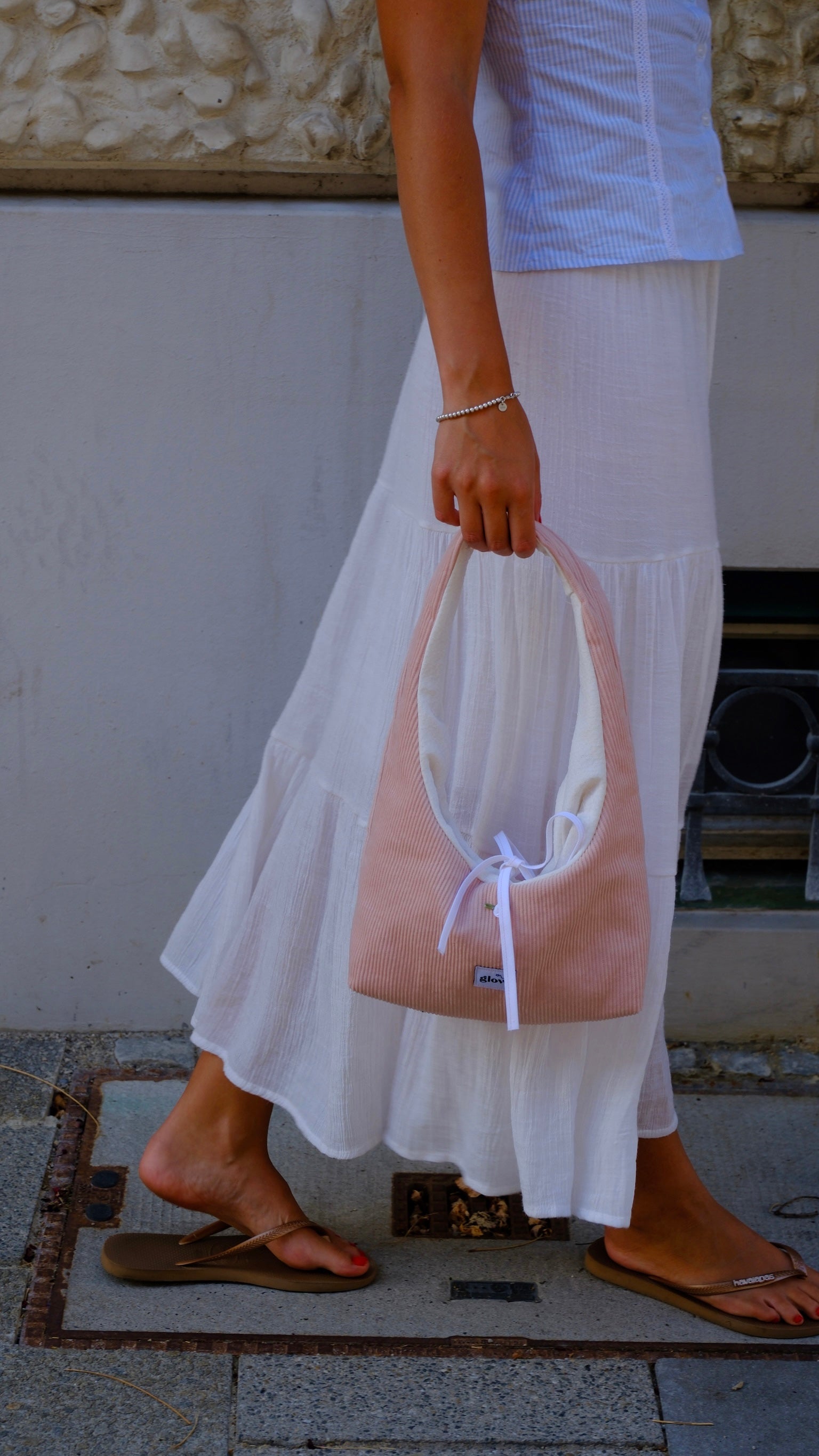 A person wearing a white tiered maxi skirt and a light blue striped top holds a dusty pink slouchy shoulder bag with a white bow detail. The person is wearing brown flip-flop sandals and standing outdoors on a stone floor.