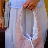 A person wearing a light blue and white striped top and white skirt holds a blush pink corduroy hobo bag with a white ribbon bow. The bag has a small white label that reads 'shoes'.