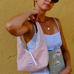 A woman wearing black sunglasses, a light blue ribbed camisole, and a white skirt carries a light pink corduroy shoulder bag with white drawstrings. She is holding a grey notebook with 'glow' written on it, set against a textured yellow wall.