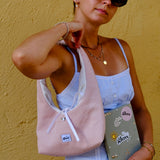 A woman wearing black sunglasses, a light blue ribbed camisole, and a white skirt carries a light pink corduroy shoulder bag with white drawstrings. She is holding a grey notebook with 'glow' written on it, set against a textured yellow wall.