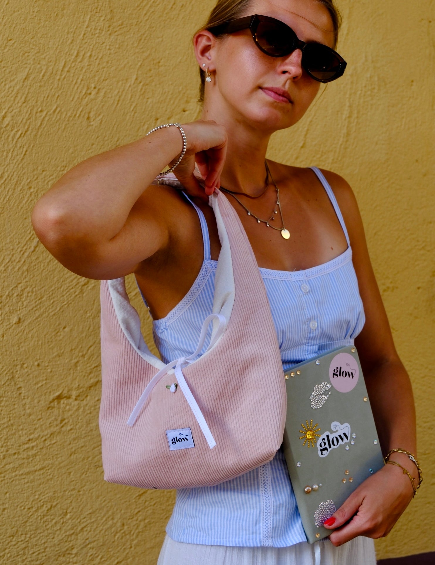 A woman wearing black sunglasses, a light blue ribbed camisole, and a white skirt carries a light pink corduroy shoulder bag with white drawstrings. She is holding a grey notebook with 'glow' written on it, set against a textured yellow wall.