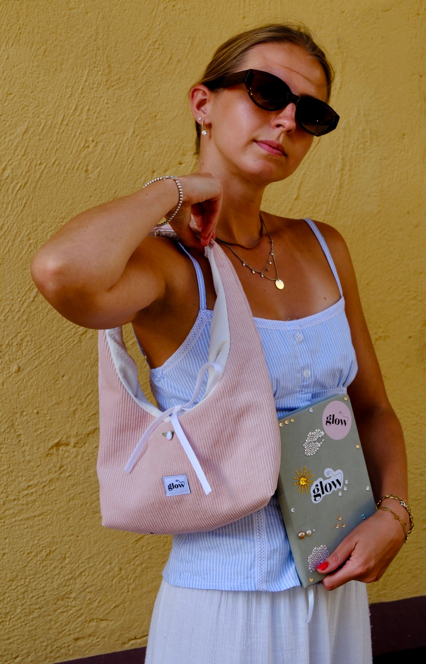 A woman wearing black sunglasses, a light blue ribbed camisole, and a white skirt carries a light pink corduroy shoulder bag with white drawstrings. She is holding a grey notebook with 'glow' written on it, set against a textured yellow wall.