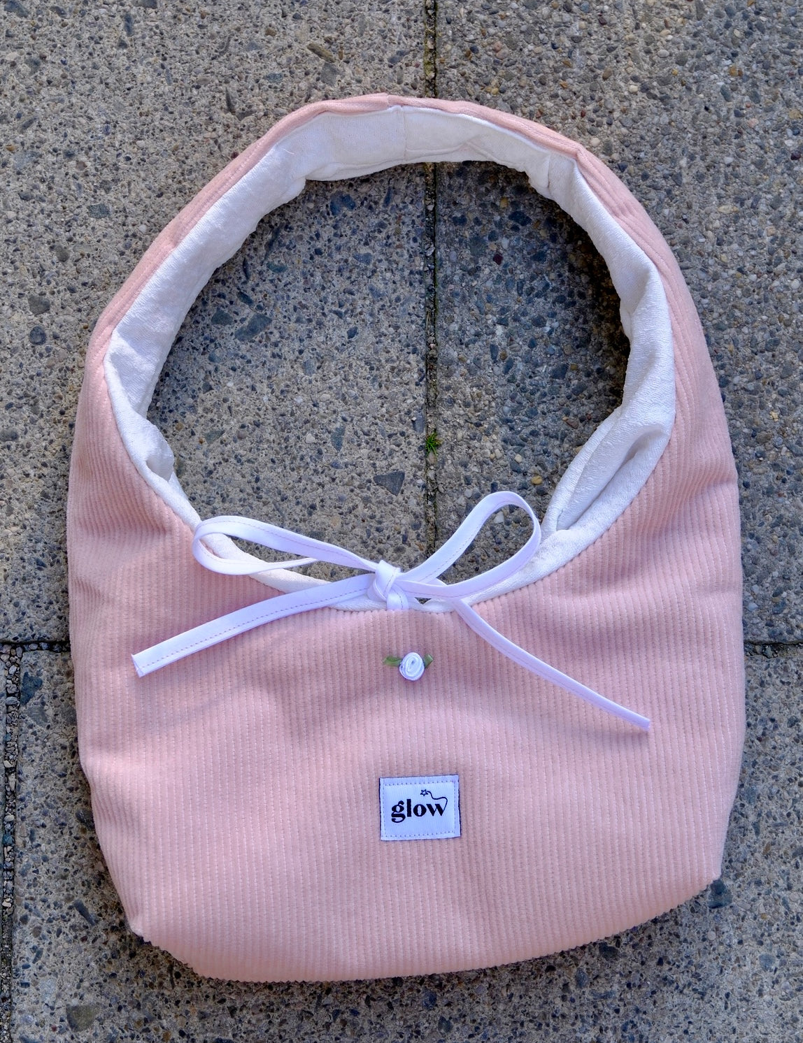 A light pink corduroy baby bib with a white inner lining, white ribbon bow, and a small white button, laid flat on a gray concrete surface. The brand label 'glow' is visible on the front.