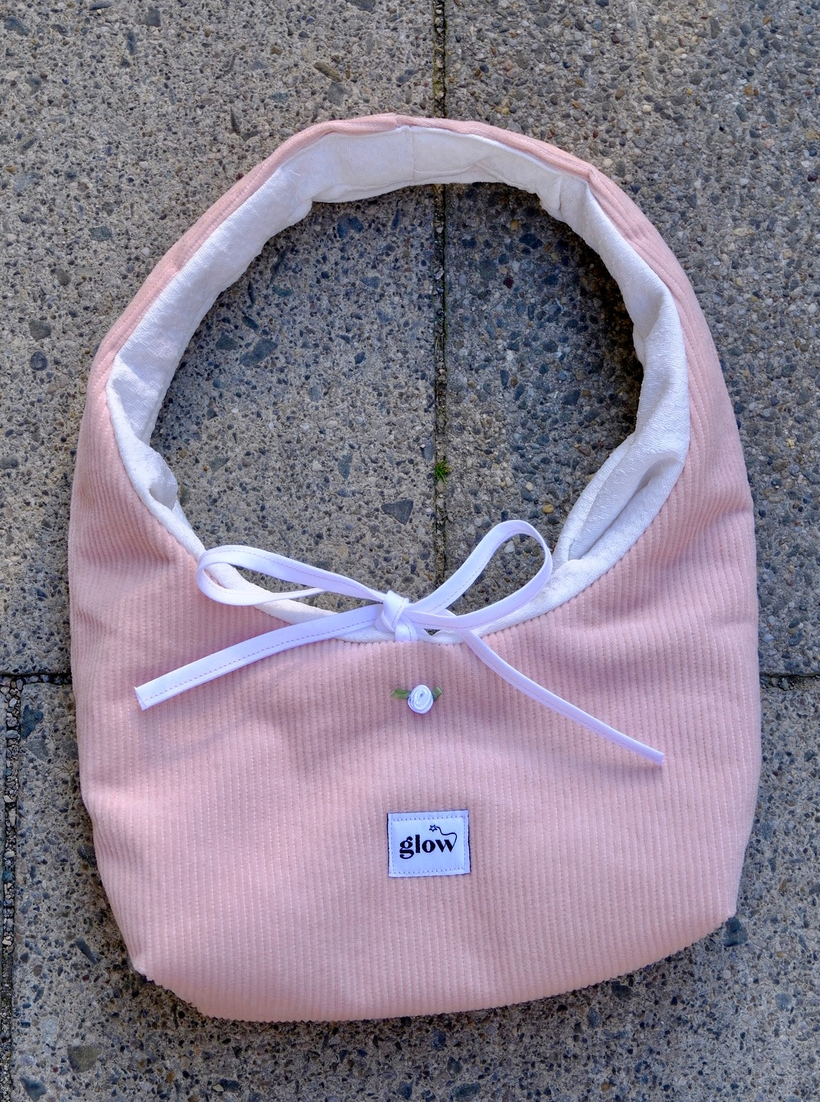 A light pink corduroy baby bib with a white inner lining, white ribbon bow, and a small white button, laid flat on a gray concrete surface. The brand label 'glow' is visible on the front.