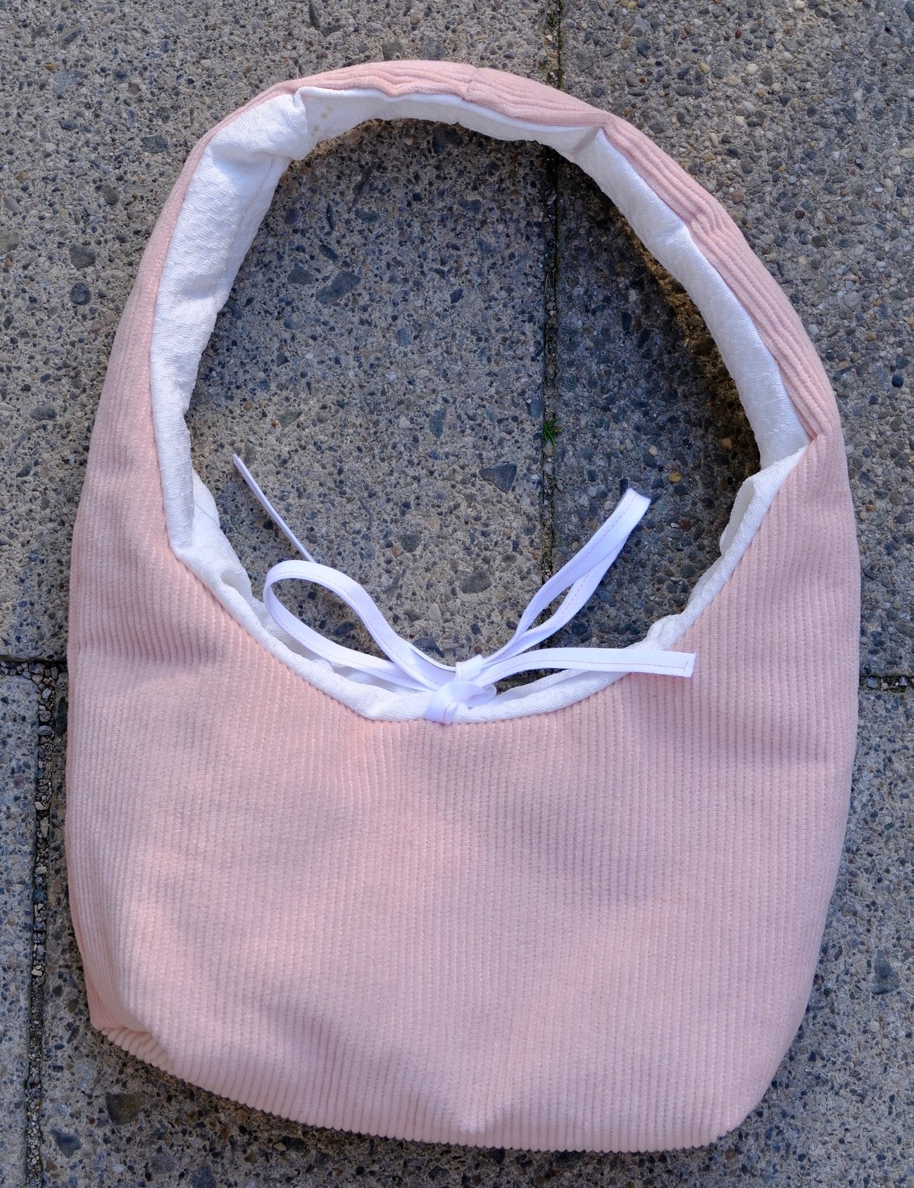 A light pink ribbed fabric headband with a white inner lining and a white ribbon bow, laid flat on a gray concrete surface.
