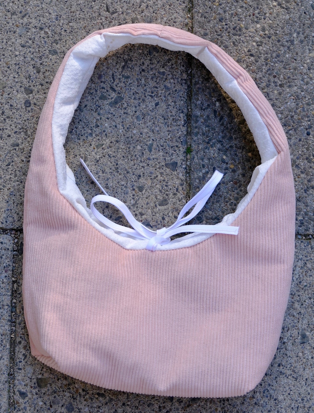 A light pink ribbed fabric headband with a white inner lining and a white ribbon bow, laid flat on a gray concrete surface.