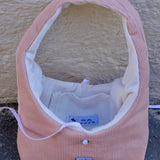 A pink corduroy pet crate or basket-style bed with a white fleece interior, photographed outdoors against a textured beige wall and gray ground. A small sewn label is visible on the front.