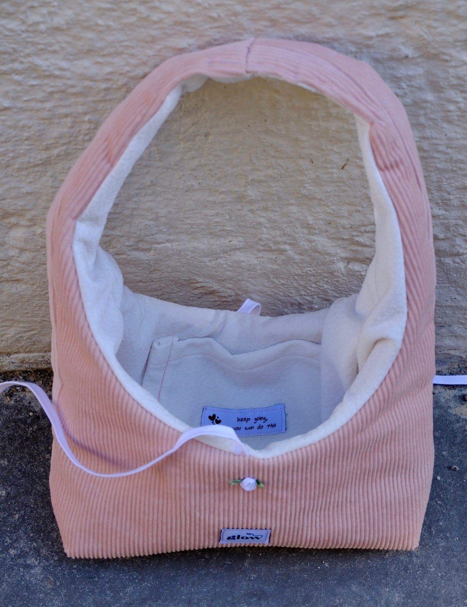A pink corduroy pet crate or basket-style bed with a white fleece interior, photographed outdoors against a textured beige wall and gray ground. A small sewn label is visible on the front.