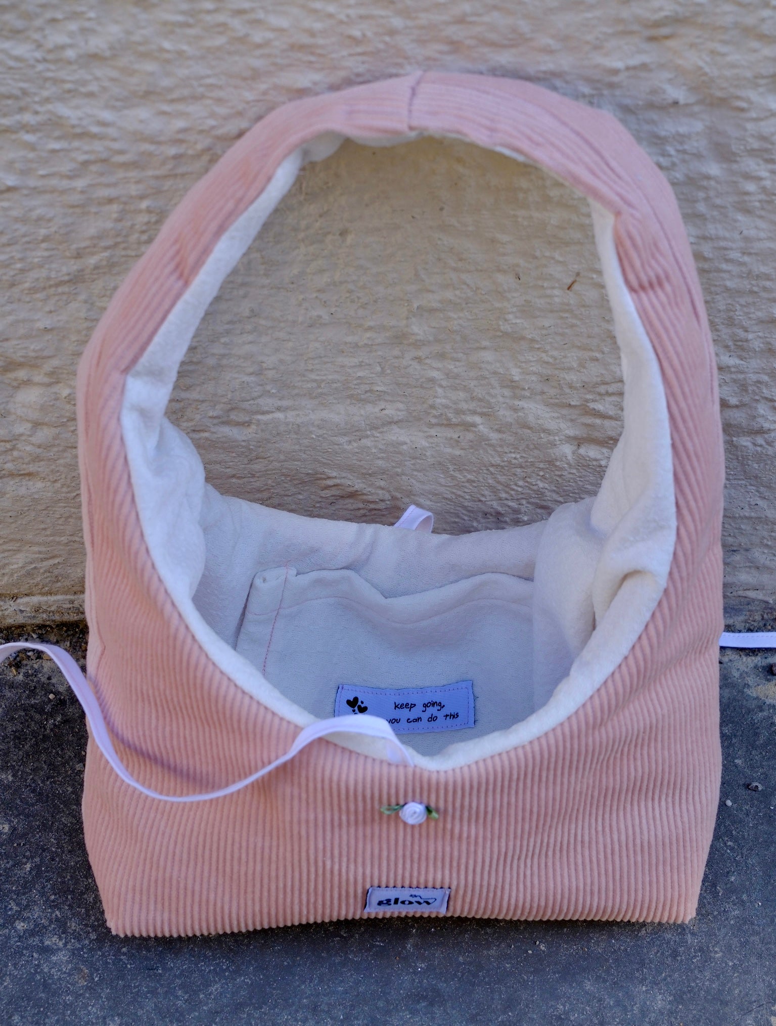 A pink corduroy pet crate or basket-style bed with a white fleece interior, photographed outdoors against a textured beige wall and gray ground. A small sewn label is visible on the front.