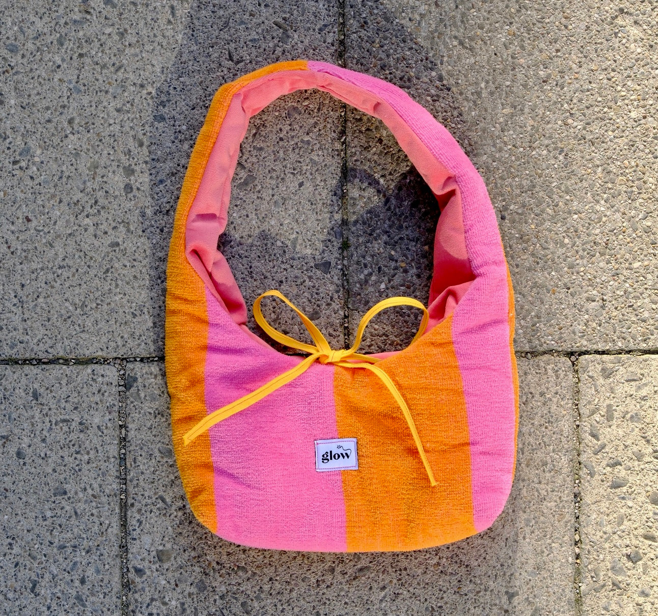 A handmade fabric shoulder bag with vertical stripes of pink, orange, and light pink, featuring a yellow ribbon bow at the top and a small white brand label that reads 'glow'. The bag is photographed from above on a gray concrete tile surface.