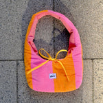 A handmade fabric shoulder bag with vertical stripes of pink, orange, and light pink, featuring a yellow ribbon bow at the top and a small white brand label that reads 'glow'. The bag is photographed from above on a gray concrete tile surface.