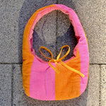 A handmade fabric shoulder bag with vertical stripes of orange and pink, featuring a yellow tie detail at the top, laid flat on gray stone tiles.