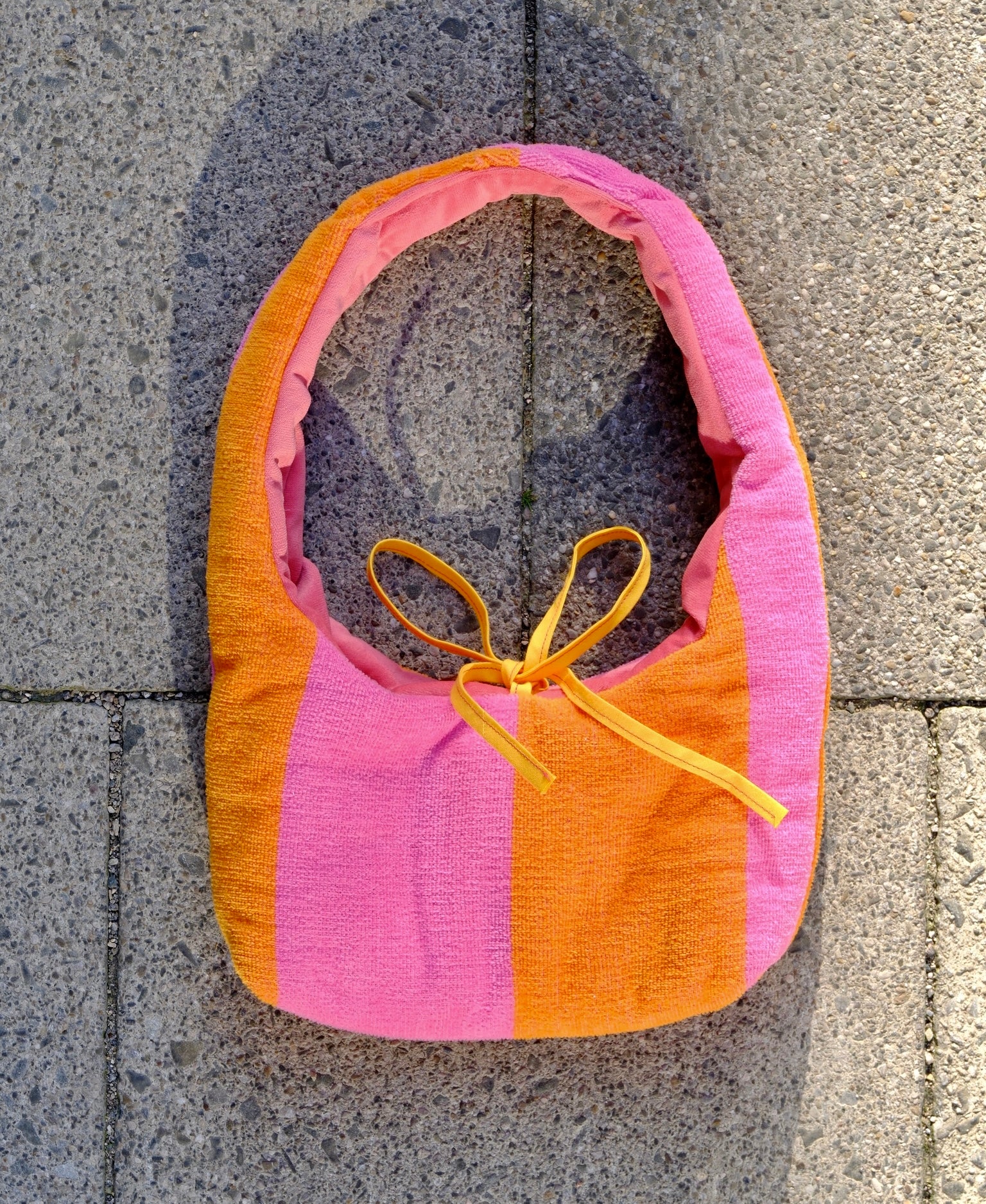A handmade fabric shoulder bag with vertical stripes of orange and pink, featuring a yellow tie detail at the top, laid flat on gray stone tiles.