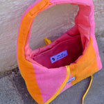 A pink and orange striped dog carrier bag made of textured fabric, shown open on a concrete surface against a stucco wall.
