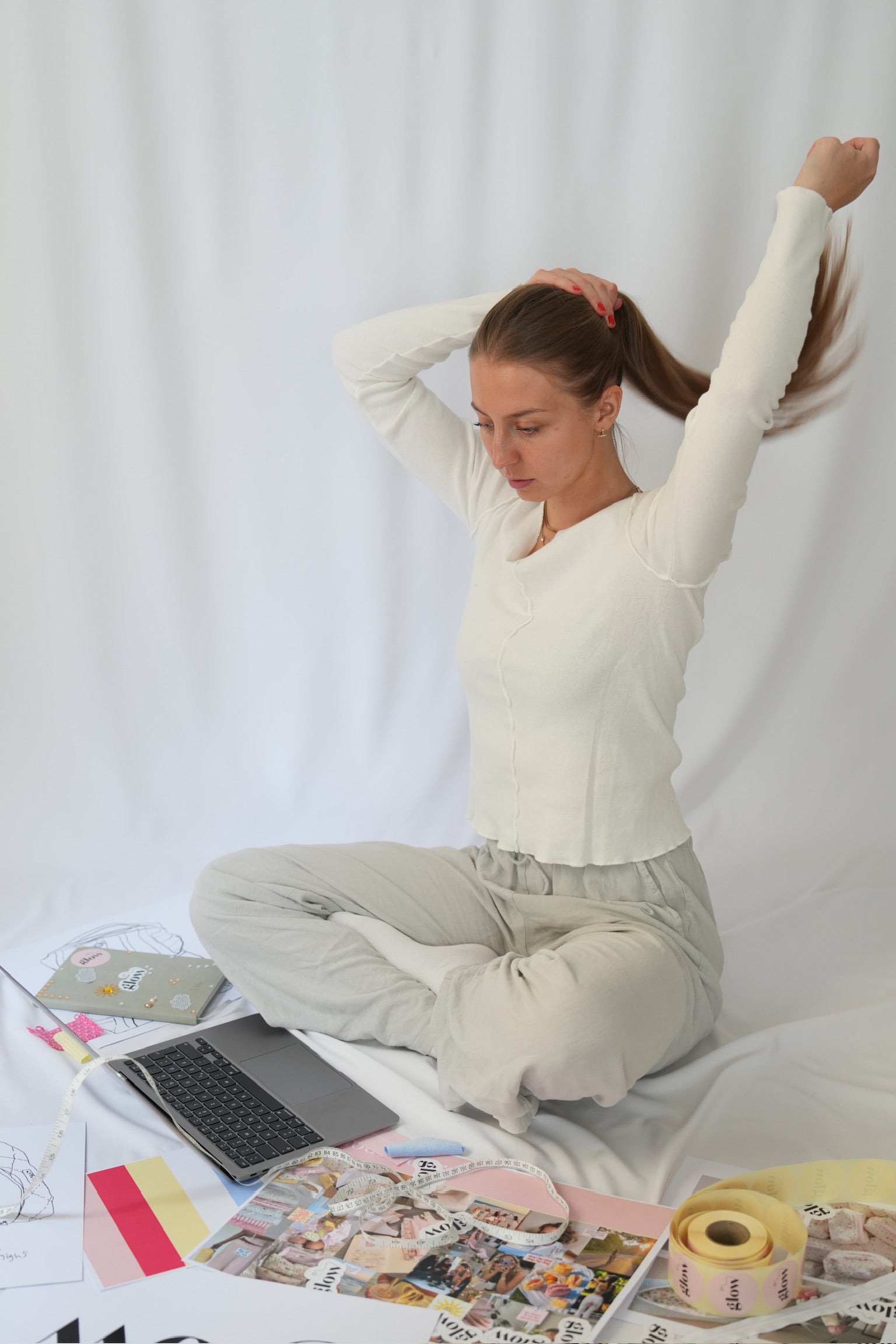 A woman sits cross-legged on a white fabric backdrop, stretching her arms overhead. She is wearing a white ribbed long-sleeve top and light beige pants.