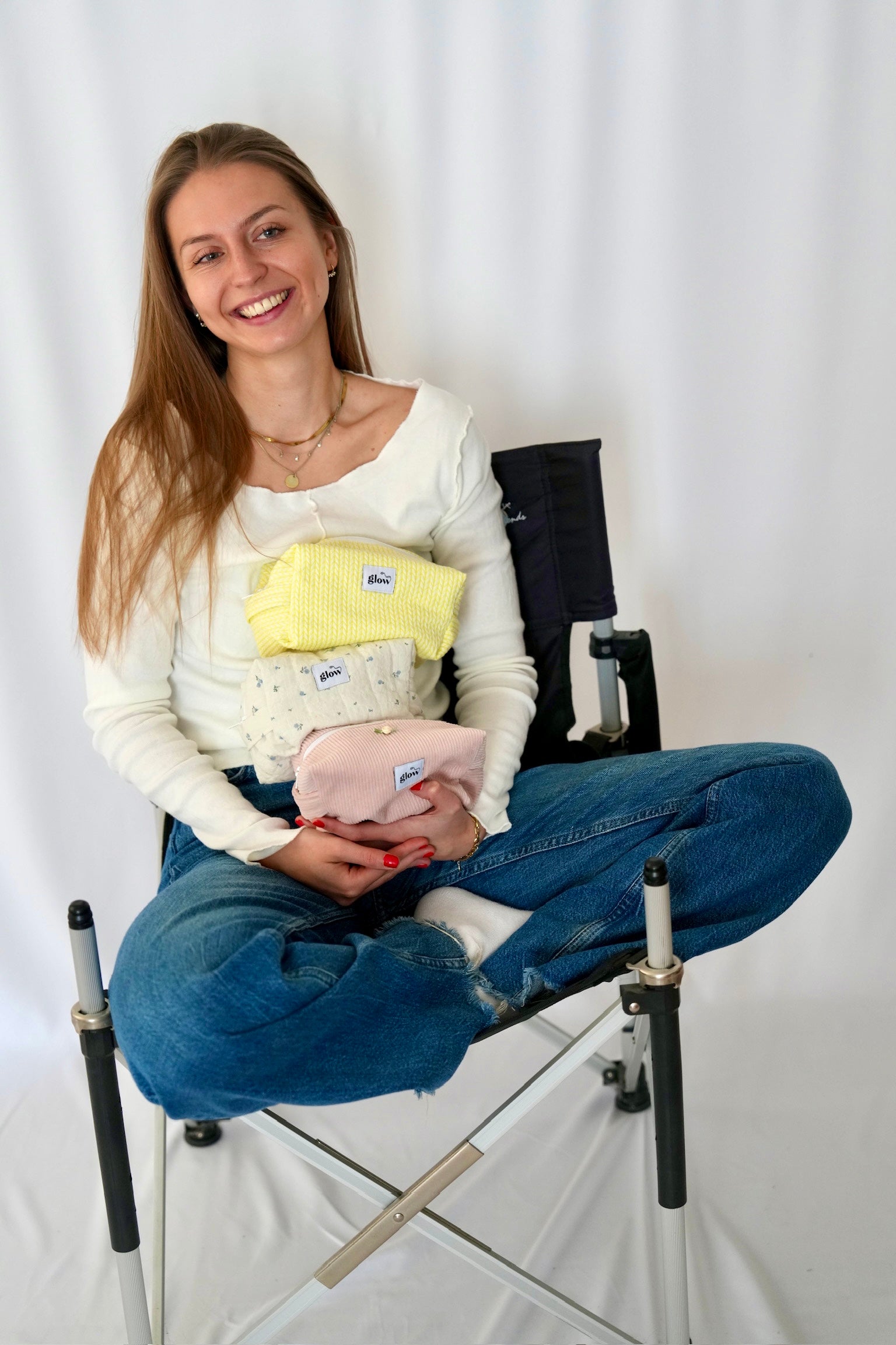 A woman sitting on a white studio chair holds three small fabric pouches or packets in different colors: yellow, white with a pattern, and light pink. Each packet has a small blue label.