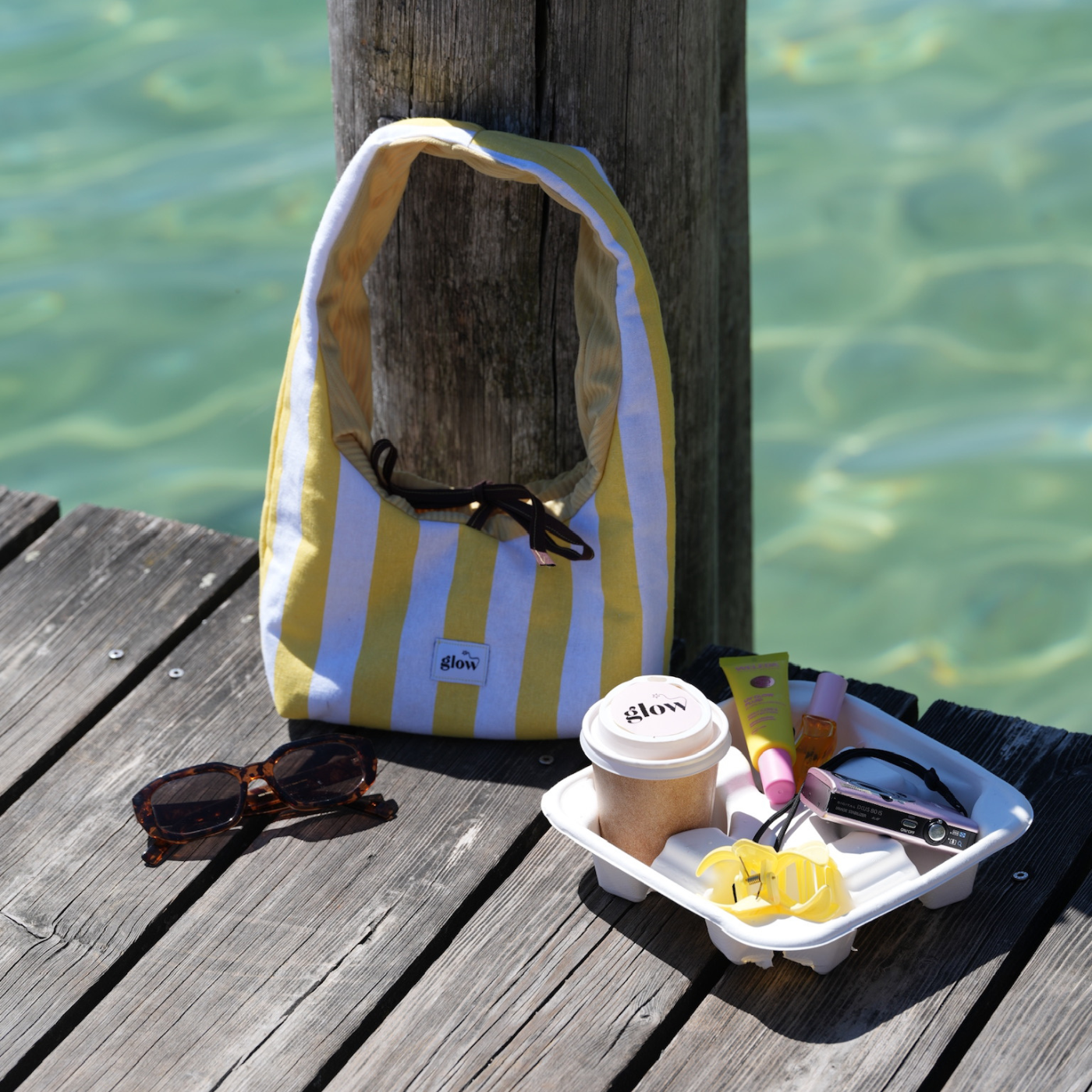 A yellow and white striped canvas tote bag with black handles sits on a weathered wooden dock next to the water. In the foreground, a white tray holds a coffee cup, sunglasses, a pink tube, and yellow items, with the bag and dock creating a beach-like outdoor setting.