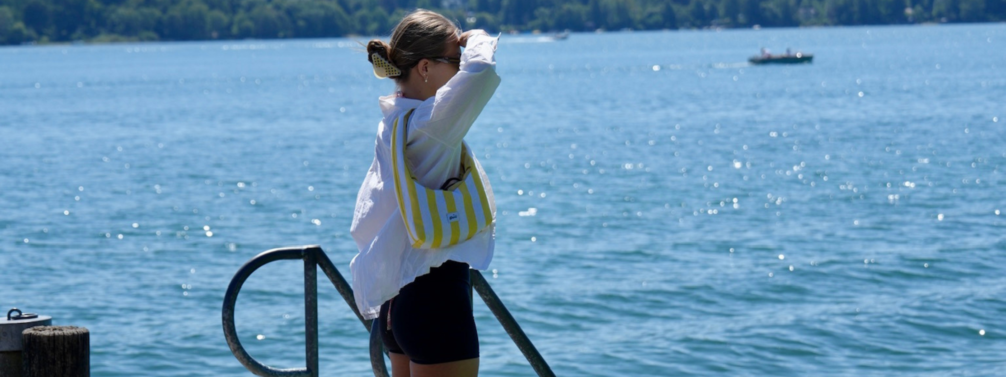 A woman standing on a dock by a large blue lake, wearing a white long-sleeve top, black shorts, and a yellow-and-white striped shoulder bag with boats and trees visible in the background.